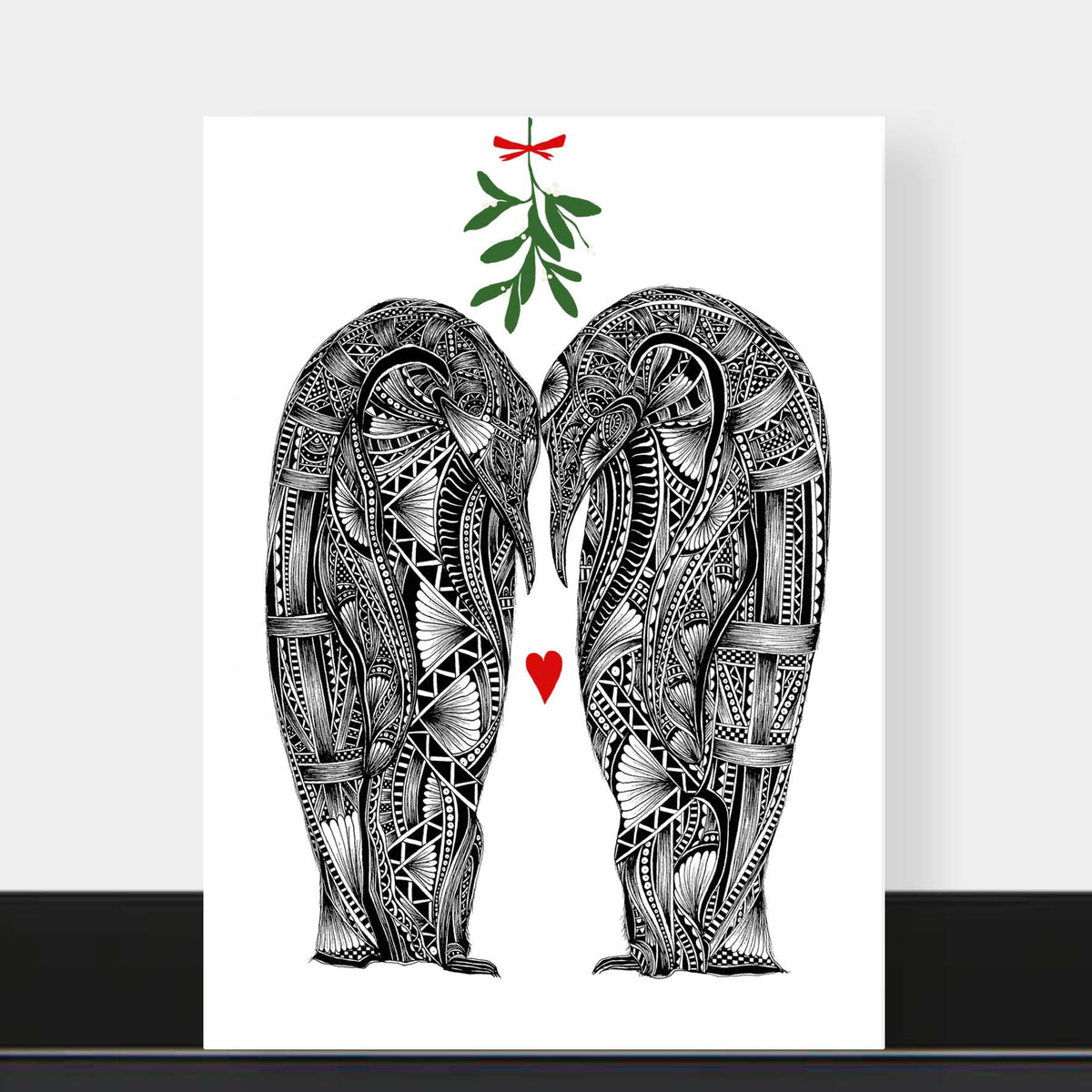 Christmas love greetings card featuring two black and white penguins with their heads together with green mistletoe above with red bow and love heart in-between them and white background. The card is on a black shelf with a grey background