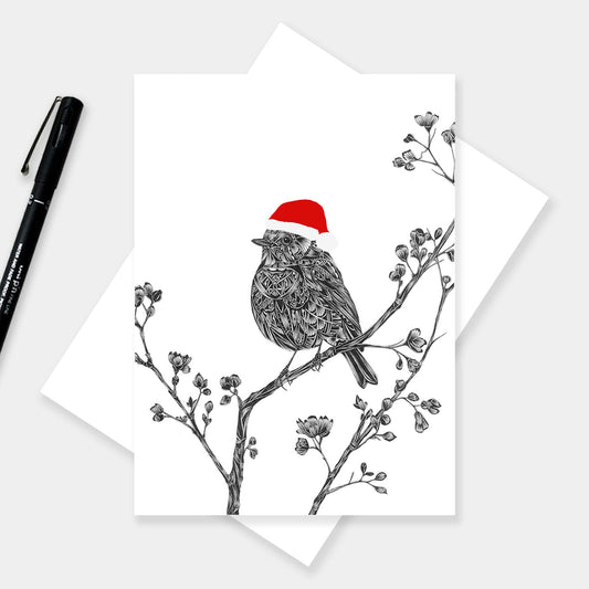 A white greeting card featuring a detailed black line art illustration of a robin on a branch with a festive red santa hat. The card is displayed on top of a white envelope, with a black pen on the left, against a light grey background.
