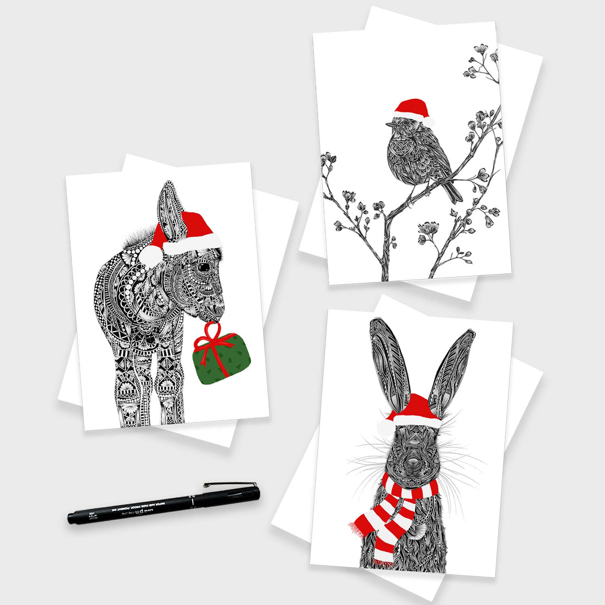 3 festive animal greetings cards featuring a robin on a branch with red Santa hat, a donkey wearing red Santa hat and green holiday present with red bow and a hare wearing a red Santa hat and scarf. The three cards are onto of white envelopes with a grey background and black pen