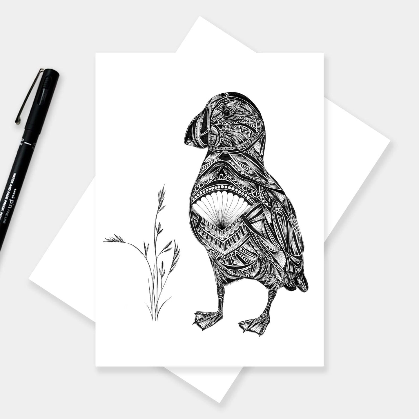 A black and white art print of a detailed puffin with intricate geometric patterns, displayed on a white card with a matching envelope and a black pen on a light gray background.