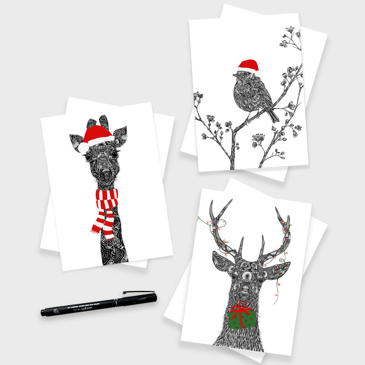 Three white cards featuring black line drawings of a giraffe in a Santa hat and scarf, a robin on a branch with a Santa hat, and a deer with antlers and a gift box, arranged on a light grey background with a black pen.