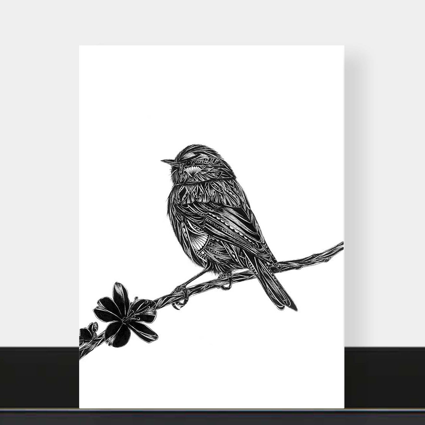 A vertical black and white art print featuring a detailed sketch of a bird perched on a branch with a flower, displayed against a light gray wall with a black baseboard.