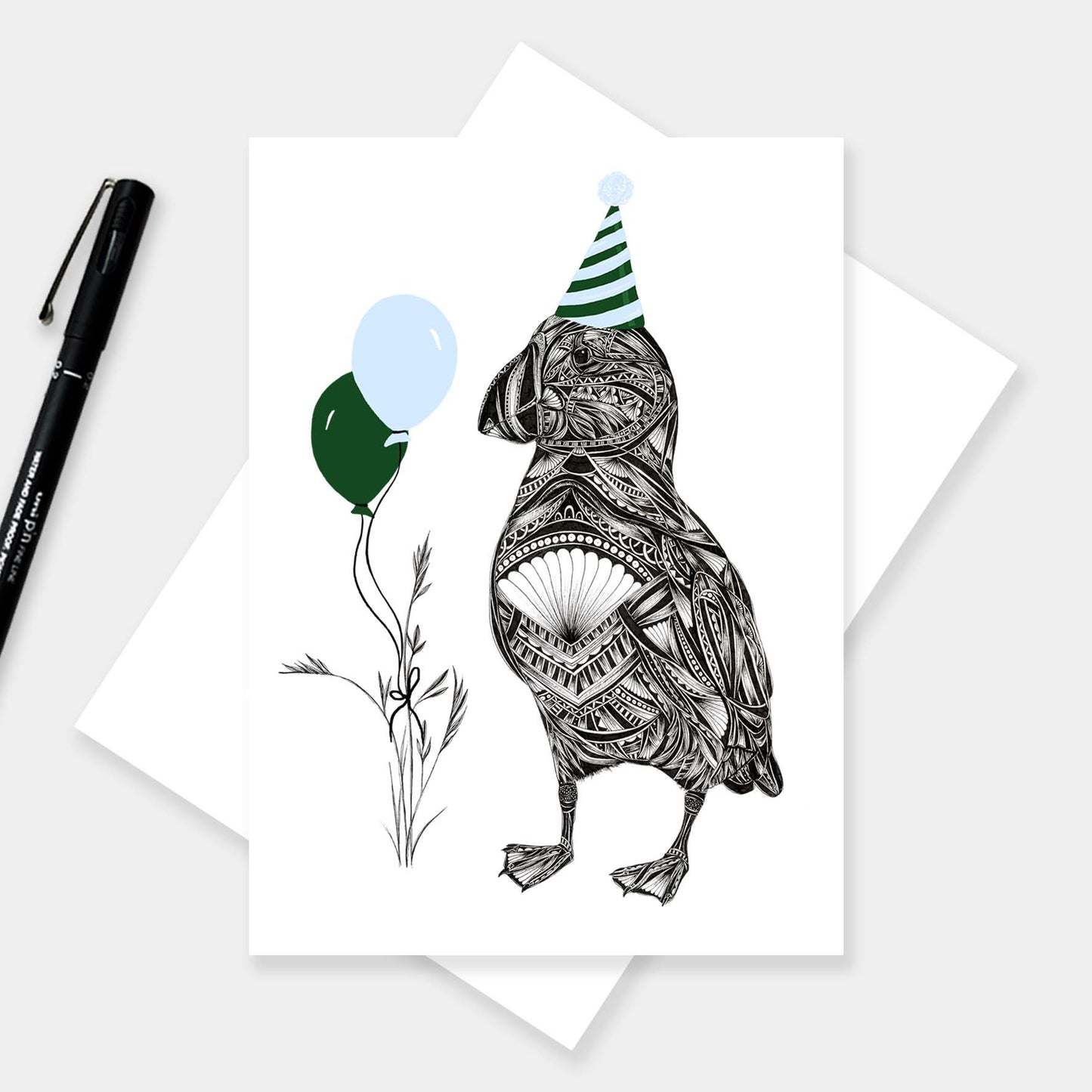 A white greeting card featuring a black line-art illustration of a puffin wearing a green and white striped party hat and holding two balloons, one dark green and one light blue. The card is displayed on top of an envelope, with a black pen on a light grey background.