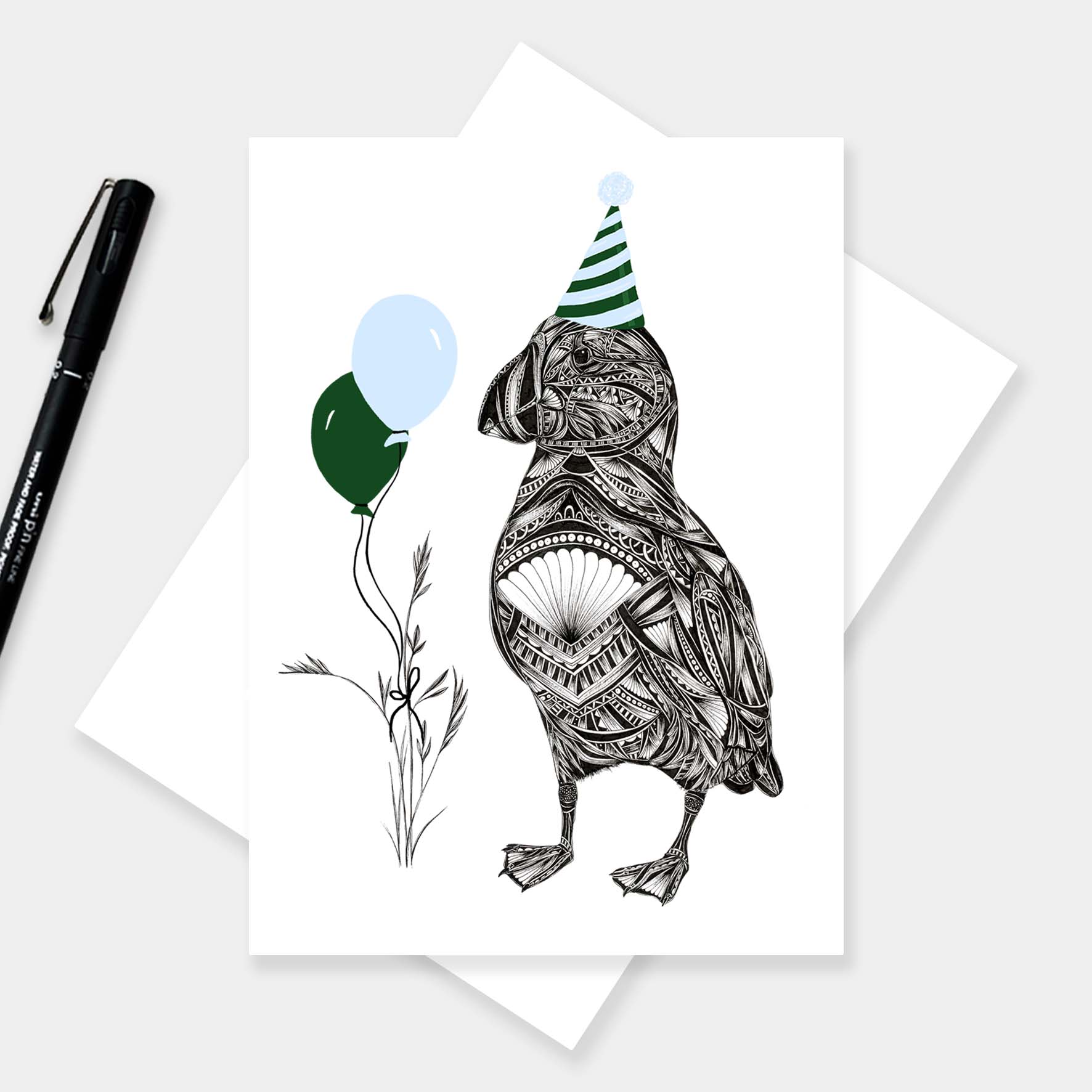 A white greeting card featuring a black line-art illustration of a puffin wearing a green and white striped party hat and holding two balloons, one dark green and one light blue. The card is displayed on top of an envelope, with a black pen on a light grey background.
