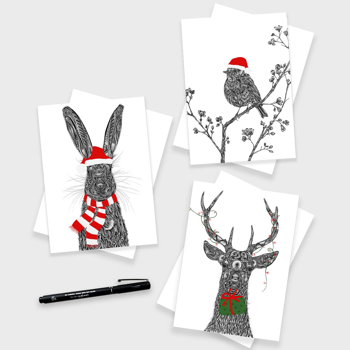 Three Christmas-themed greetings cards with black and white animal illustrations are arranged on a light grey background with a black pen. The prints show a rabbit in a red Santa hat and striped scarf, a robin on a branch wearing a red Santa hat, and a deer head with antlers wearing a red and green gift box.