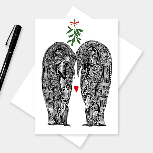 Christmas love greetings card featuring two black and white penguins with their heads together with green mistletoe above with red bow and love heart in-between them and white background. The card is on a white envelope with grey background and black pen