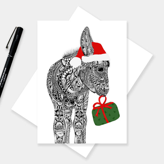 Christmas card featuring a donkey illustration wearing a Santa hat and a green holiday gift with red bow with a white background. The card is on a white envelope with a grey background and black pen