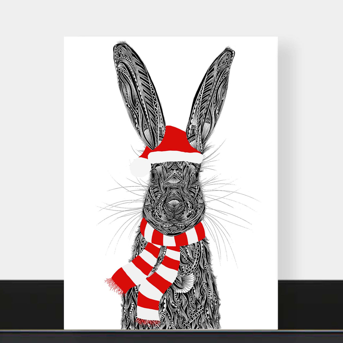 Christmas card featuring hare illustration wearing red festive scarf and Santa hat, The card is on a black shelf with a grey background