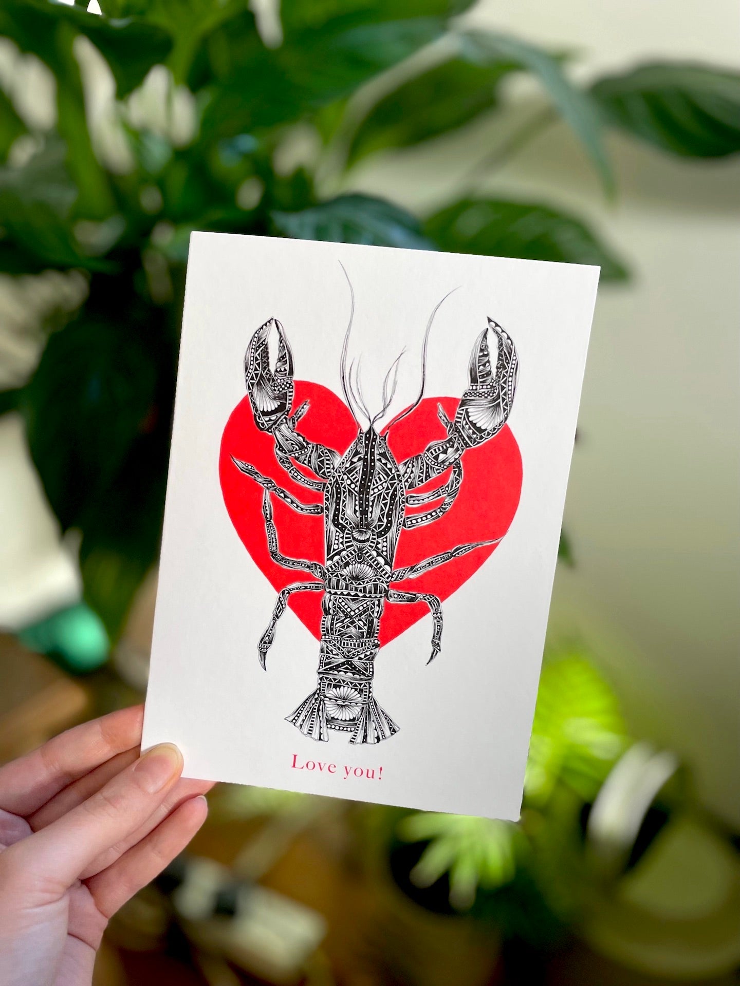 Card with a lobster design on a red heart background held by a hand with a blurred green plant background.