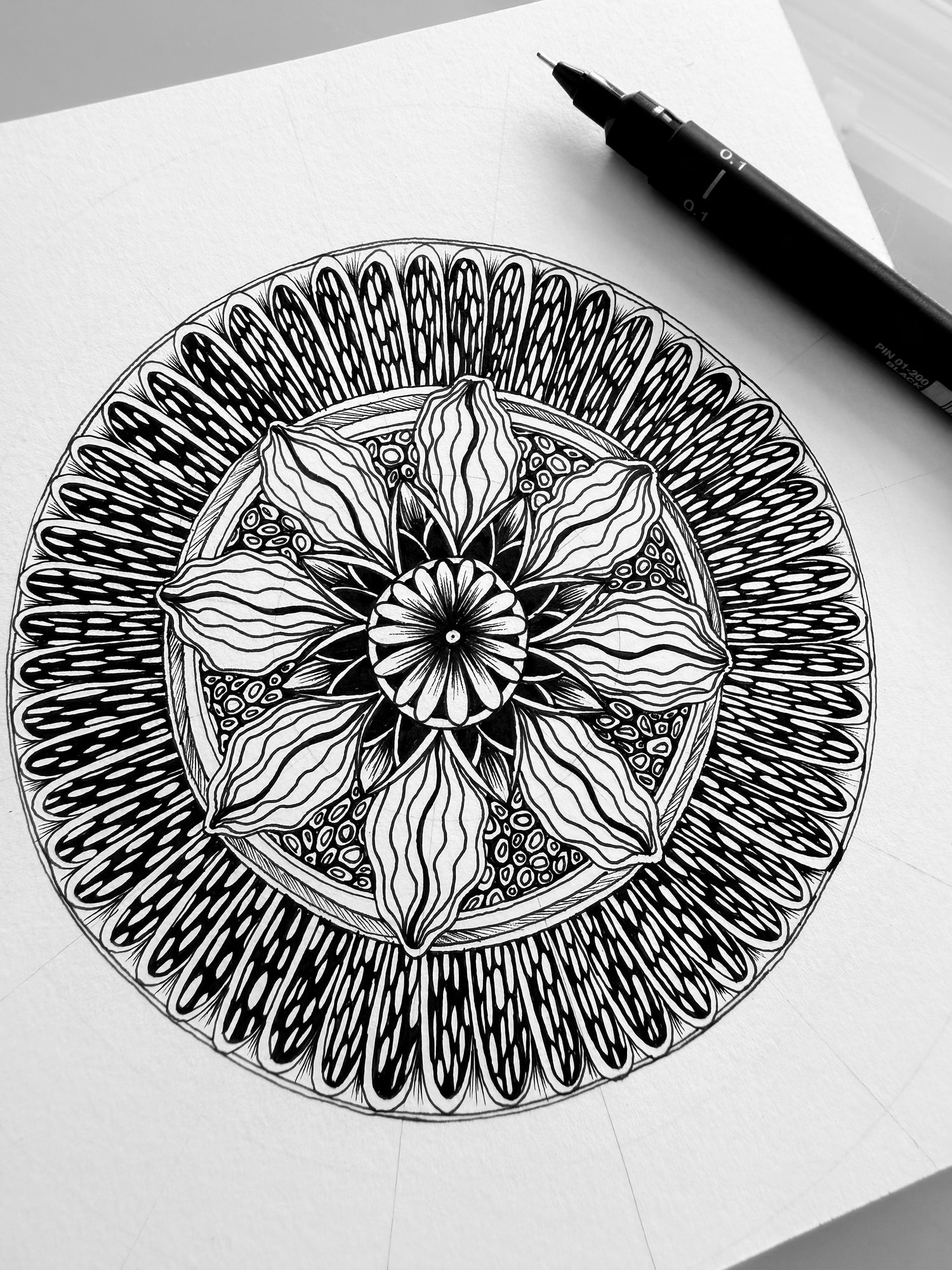 Black and white mandala drawing on a white sheet of paper with a pen next to it.