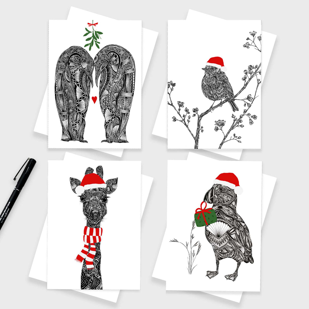 4 Christmas cards on white envelopes on grey background with black pen. The greetings cards feature 2 penguins with green mistletoe and red love heart, a robin on a branch wearing a red Santa hat, a giraffe wearing a red Santa hat and scarf and a puffin wearing a red Santa hat and green present with red bow.
