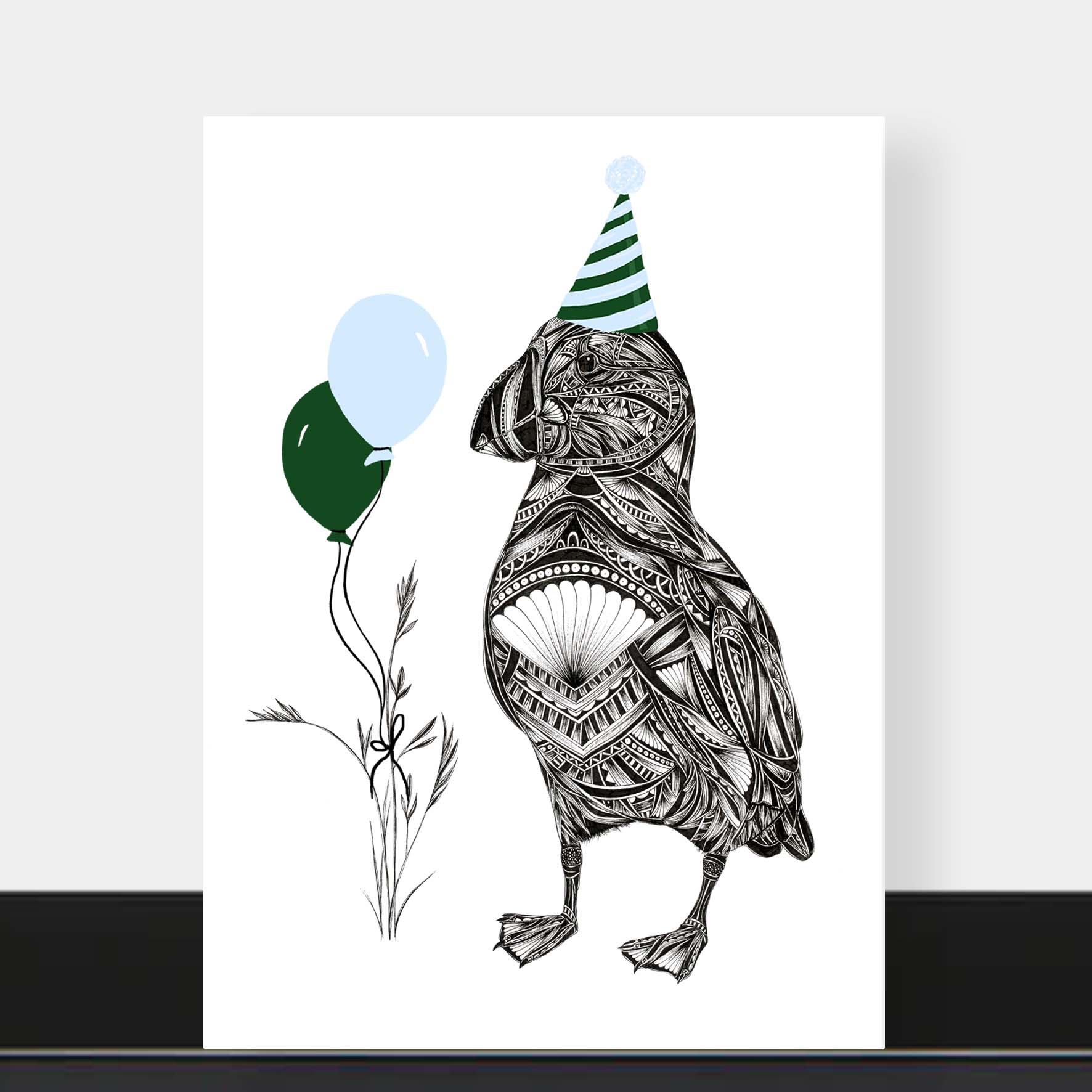 Illustration of puffin with pattern detail wearing blue and green hat with balloons in grass on white background. The card it placed on a black shelf with a grey wall in background.