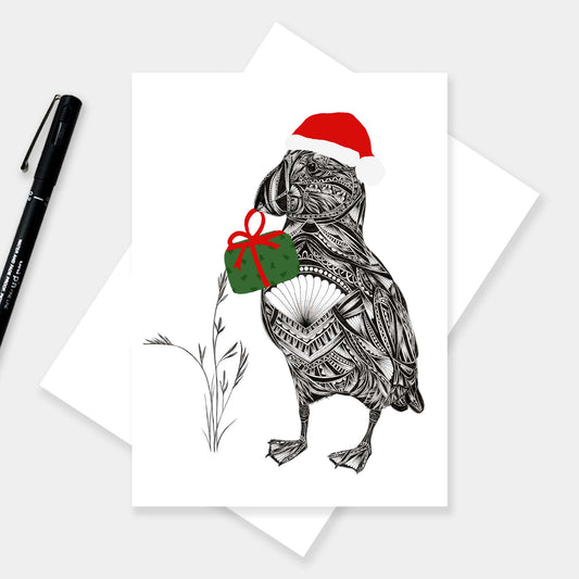 Christmas card of black and white puffin illustration holding a festive green holiday present with red bow, wearing a red Santa hat. The card is onto of a white envelope on a grey background with black pen to the side.