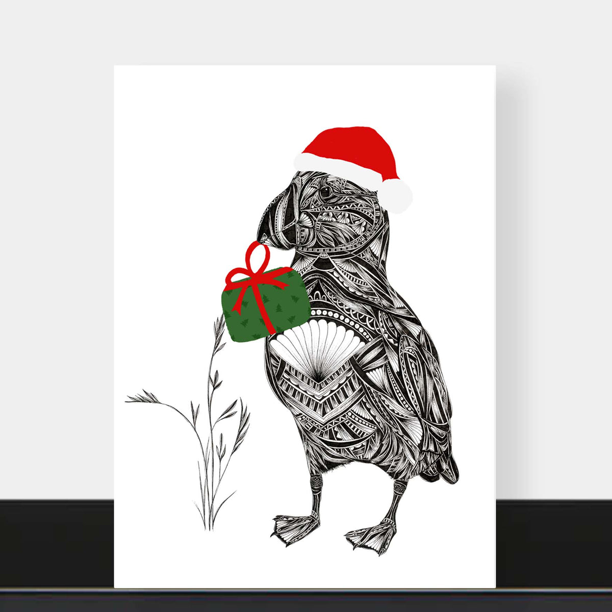greetings card of puffin illustration in black and white holding a festive green holiday present with red bow, and wearing a red Santa hat. The card is on a grey background on a black shelf