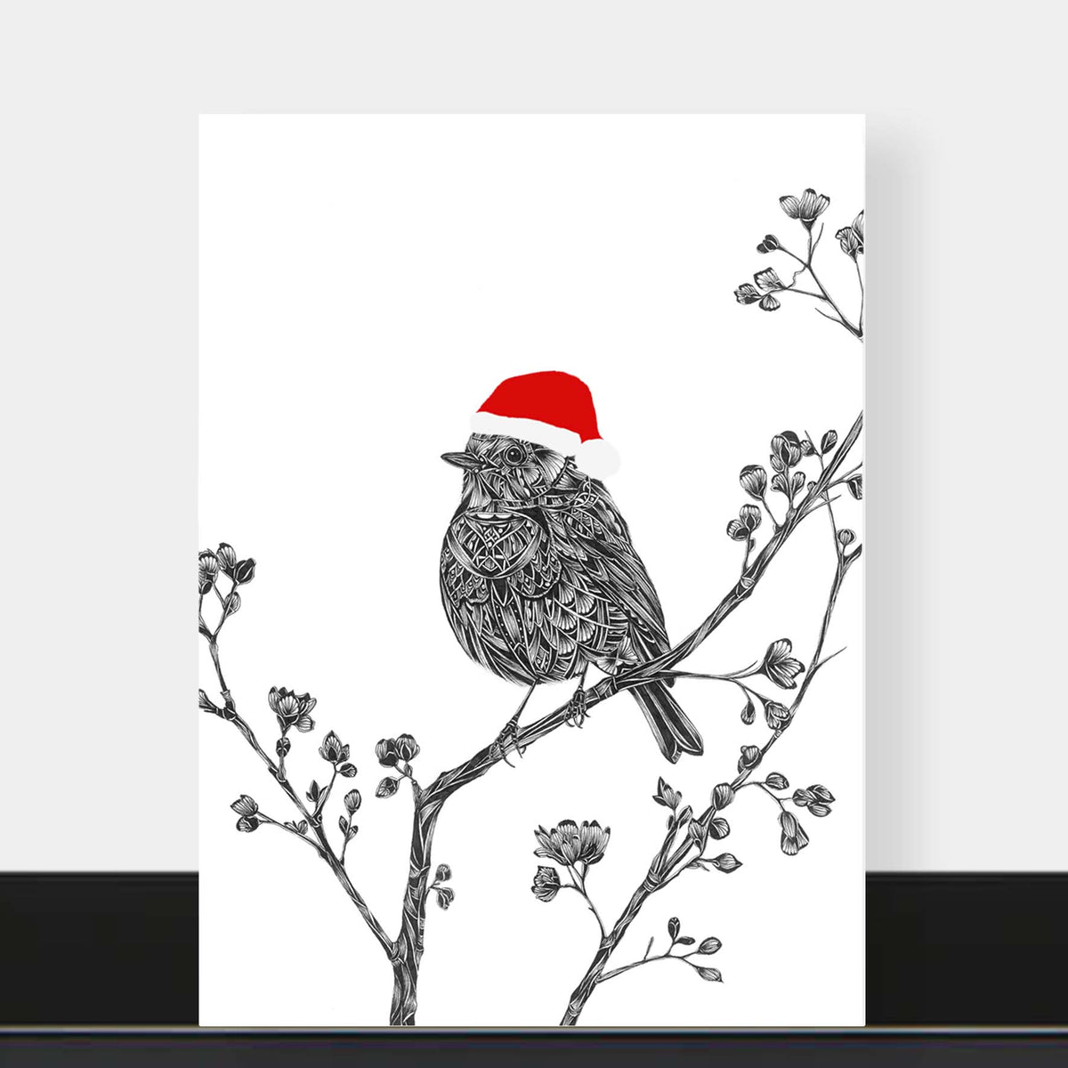 A vertical greetings card featuring a black and white sketch of a robin perched on flowering branches, wearing a red Santa hat, displayed against a light grey wall above a dark surface.