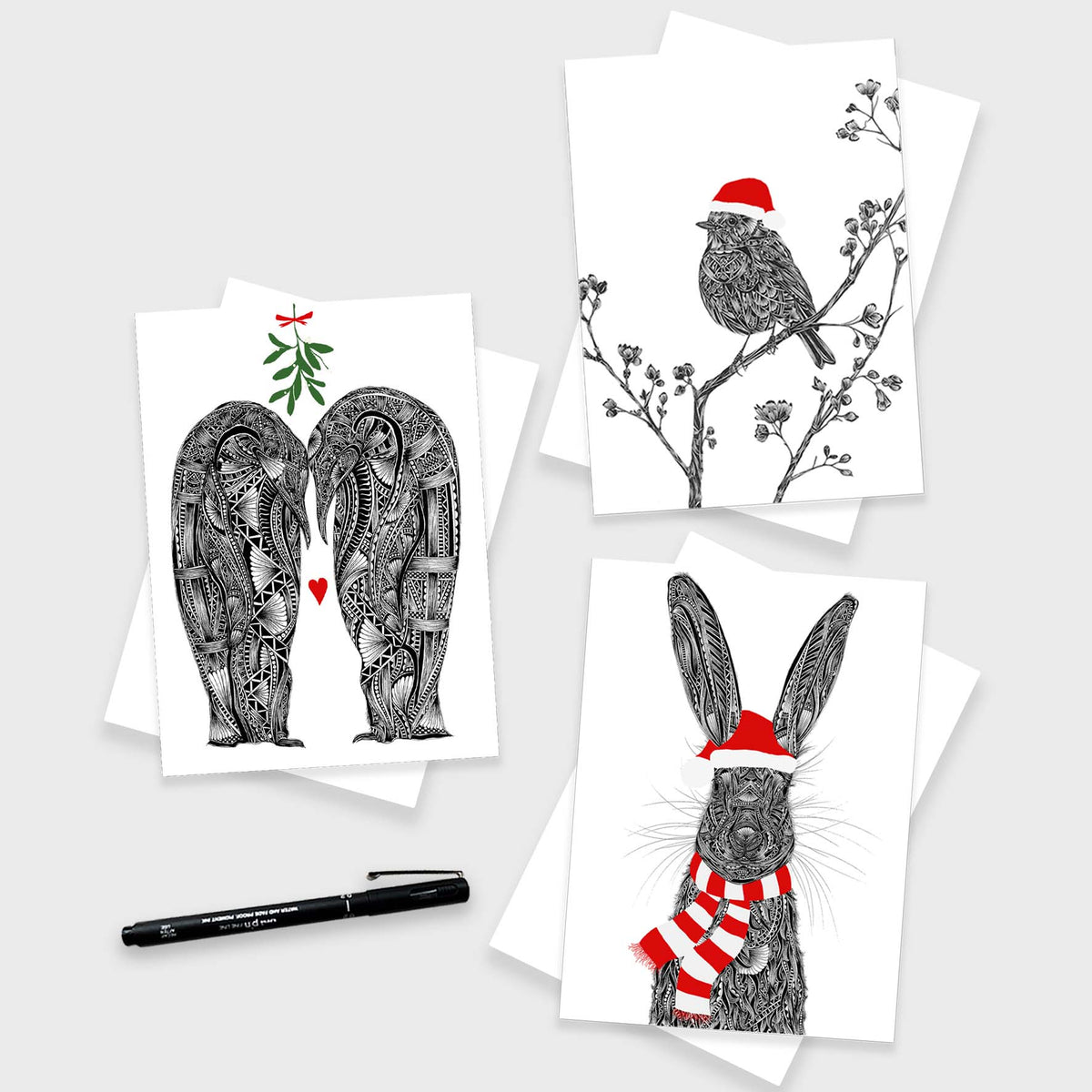 three Christmas cards on white envelopes with grey background and pen. The cards feature penguins with green mistletoe above them and love heart in-between them, a robin on a branch with a red Santa hat and a hare with red Santa hat and scarf.