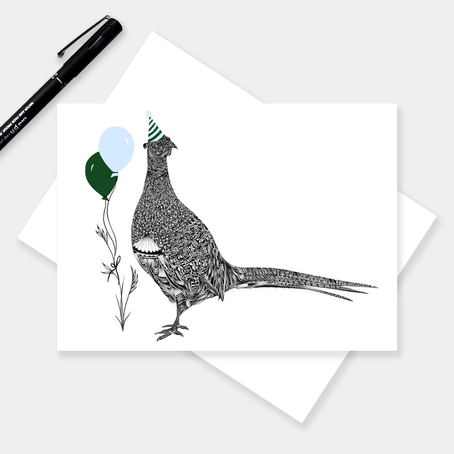 A horizontal greeting card with a black-and-white illustrated pheasant wearing a party hat and holding balloons, shown on top of a white envelope. A black pen rests in the upper left on a light grey background.