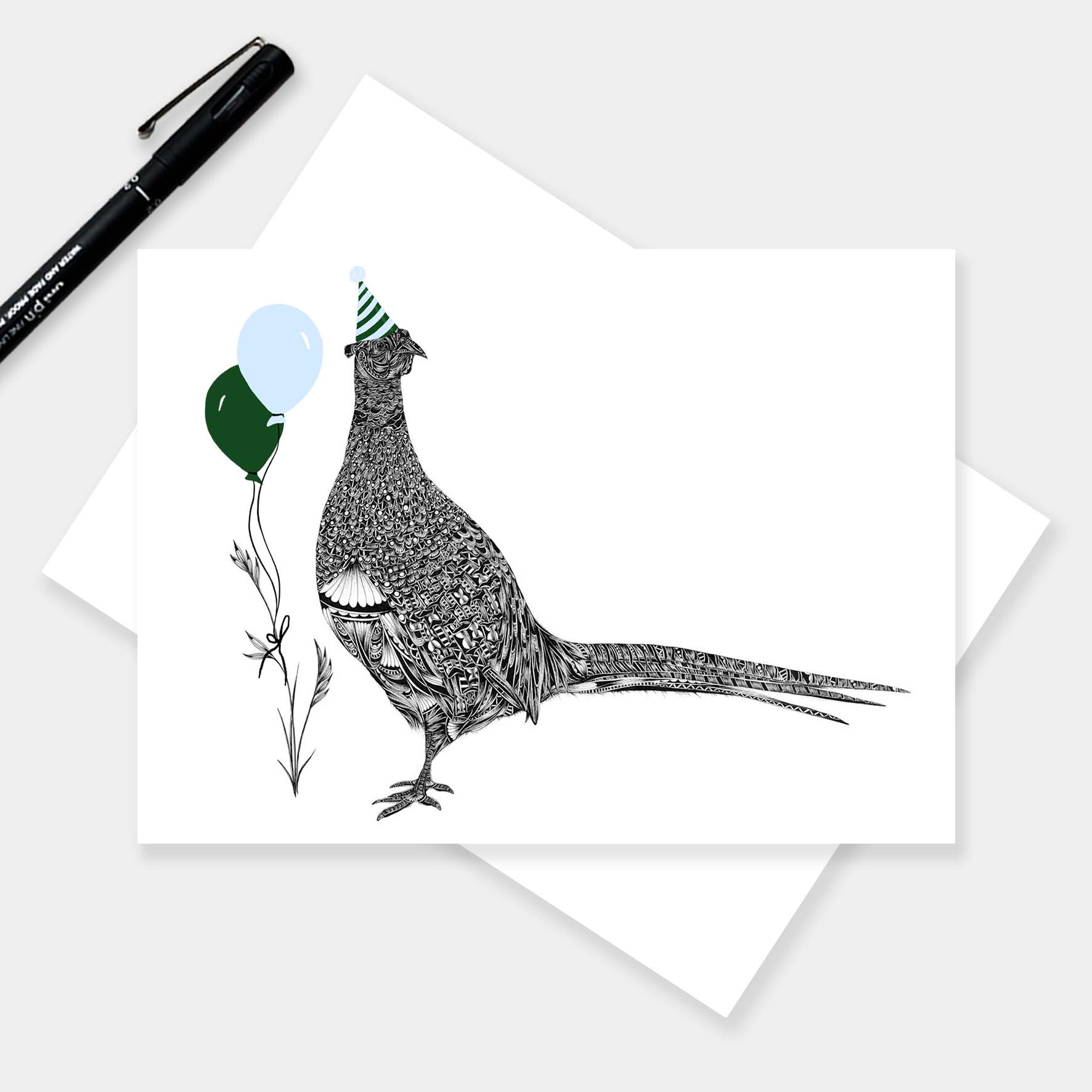 A horizontal greeting card with a black-and-white illustrated pheasant wearing a party hat and holding balloons, shown on top of a white envelope. A black pen rests in the upper left on a light grey background.