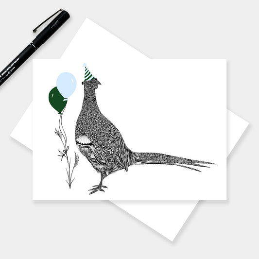 A horizontal greeting card with a black-and-white illustrated pheasant wearing a party hat and holding balloons, shown on top of a white envelope. A black pen rests in the upper left on a light grey background.