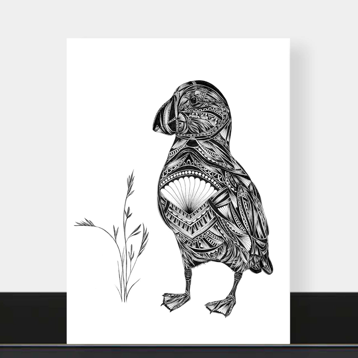 A black and white art print featuring a detailed illustration of a puffin filled with intricate tribal patterns, standing next to a small sprig of grass. The print is displayed against a light gray wall with a black baseboard visible at the bottom.