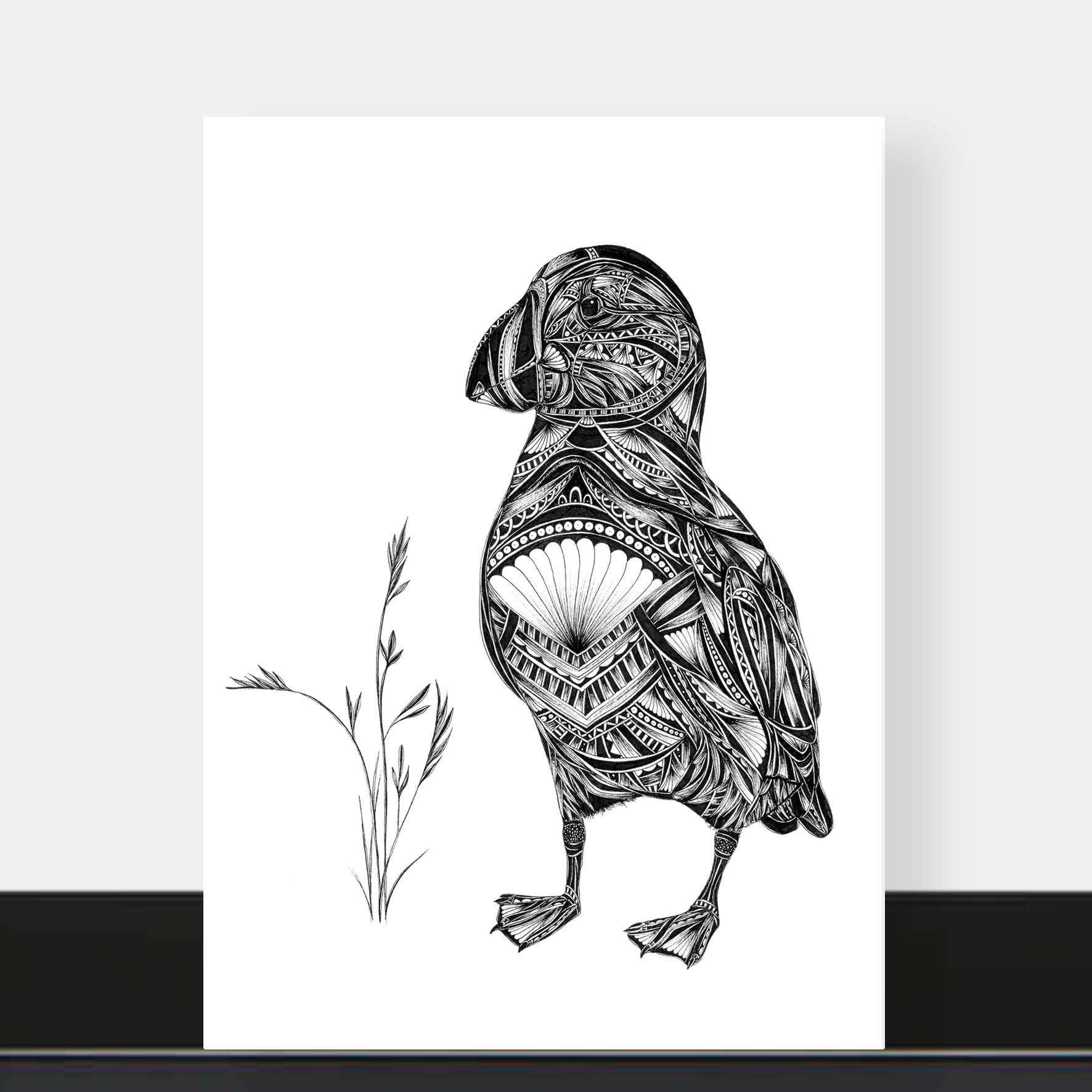 A black and white art print featuring a detailed illustration of a puffin filled with intricate tribal patterns, standing next to a small sprig of grass. The print is displayed against a light gray wall with a black baseboard visible at the bottom.