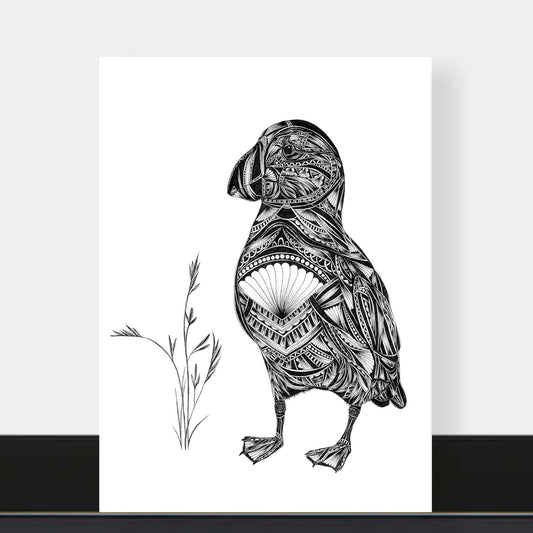 A black and white art print featuring a detailed illustration of a puffin filled with intricate tribal patterns, standing next to a small sprig of grass. The print is displayed against a light gray wall with a black baseboard visible at the bottom.