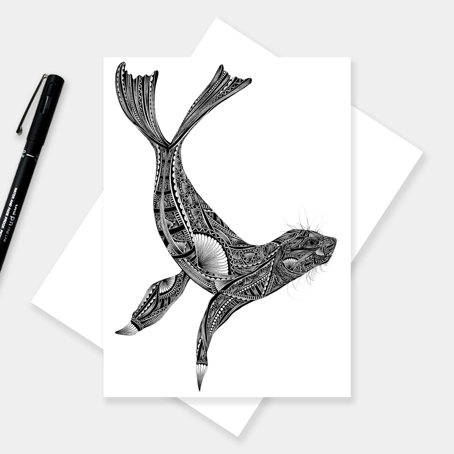 A black and white illustration of a seal with intricate geometric patterns, presented on a white card with a black pen and white envelope on a light gray background.